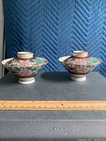 Front view of two Japanese lidded bowls with intricate floral and geometric multicolor pattern and gold bands placed on a gray surface with ruler.