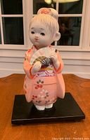 Front view of the ceramic figurine showing the girl holding a fan wearing a pink kimono with floral patterns, on a black stand.