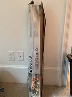 Jenn-Air Rotisserie Kit boxed and leaning against a wall, showing full length of the package with brand and kit details.