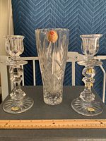 Pair of clear lead crystal candlesticks and a lead crystal vase resting on a table with a blue padded backdrop. Each item has labels indicating authenticity.