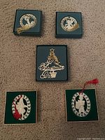 Five ornaments in green boxes arranged on carpet: two circular with gold tassels, two oval with red tassels, and one Christmas tree shape with gold bird and star.