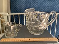 Front view of two clear pressed glass pitchers, smaller to the left and larger to the right on a black surface with ruler for scale