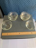 Top-down view of six small clear glass bowls arranged on a surface next to a ruler for size reference. Each bowl shows some surface smudges.