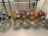 Row of 5 Smurf themed glasses showing characters Handy, Grouchy, Baker, Hefty, and Clumsy with vibrant cartoon illustrations on clear glass.