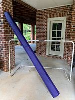 White metal soccer goal with net and blue padded balance beam mat propped against it outdoors on a concrete porch