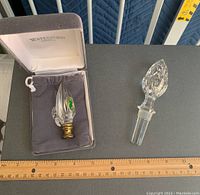 Two Waterford Crystal items side by side with measuring ruler below for scale: lamp finial with brass base and acorn-shaped bottle stopper with glass stopper.