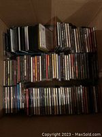 Box filled with assorted CD jewel cases stacked in rows showing various titles and plastic wraps on some cases.