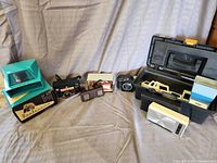 Lot of vintage electronics including cameras, slide viewers, and Hitachi radio with cases and accessories shown on cloth surface.