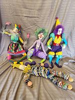 Five hand stitched clown and jester dolls in various colorful costumes posing sitting and laying on a cloth background.