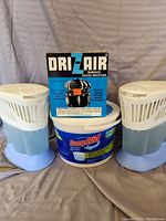 Two Dri-Z-Air moisture removers with removable tops flanking a Damp Rid moisture absorber container and a box of Dri-Z-Air moisture remover.
