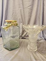 Both hexagonal glass jar with cork lid and decorative twine, and a heavy cut glass vase are shown, well lit and in front of a gray curtain backdrop.