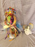 Full view of two handcrafted dolls, a colorful sorceress and a white fairy, seated with fabric backdrop