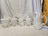 Photo showing four leaded crystal cannisters with lids and one matching pitcher against a plain cloth backdrop.