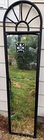 Photo of a tall, narrow rectangular mirror with a black metal frame and an arched top featuring radial spokes. Mirror is standing vertically outdoors reflecting grass and garden.