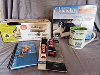 Photo showing the Neater Feeder box on the left, Snoozer Ortho Air pet bed box on the right, two dog collars in the center, and a blue notebook with a dog photo on the cover at the bottom left.