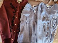 Three sweaters laid out, including one burgundy Crystal Kobe cardigan with black embroidered floral design and two blue floral cardigans