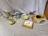 Wide view of three decorative ceramic and glass items with beach and ocean themes including seashell planter, dish with shell soaps, and orca bowl along with starfish and glass jar with seashell-topped cotton swabs.