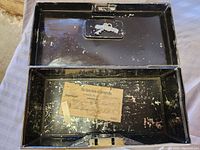 Open view of the rectangular black metal bank box showing wear and paint chips inside and outside, keys attached inside lid, faded bank slip inside base.