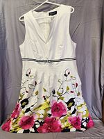 White sleeveless summer dress with floral print hanging on hanger, showing front details including v neckline and belt.
