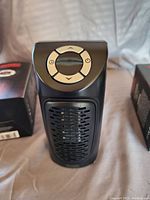 Front view of the black compact electric heater with top control panel and buttons, showing unobstructed detail of the unit and the boxes behind it.