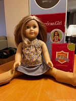 Photo of the American Girl doll seated on a wooden surface with long brown hair, floral blouse, gray skirt, and tan tights. Original box and guide book placed behind the doll.
