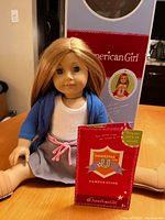 American Girl doll sitting with legs outstretched, wearing casual clothes, with original packaging and Campus Guide booklet in the background.