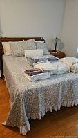 Bed made with grey paisley patterned bedspread with lace trim, white lace bed skirt, various linens packaged on top of the bed.