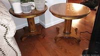 Pair of round wooden end tables shown together on hardwood floor. One table holds various small containers but items not included in lot.