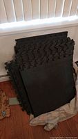 Stack of black foam floor mats with interlocking edges, some cut, folded over cloth on wooden floor near window blind.