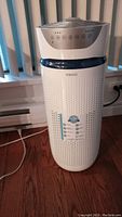 Full front view of white Homedics air purifier on hardwood floor next to window blinds with cord visible.