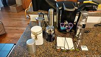 Photo showing all items together including the Keurig coffee maker, can opener, coffee grinder, thermos, napkin holder, ceramic canister, and teapots arranged on kitchen countertop.