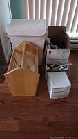 Photo showing the full lot including the cloth covered laundry hamper, wooden magazine rack, box of thread spools, and small plastic storage drawers on a wooden floor near a window with vertical blinds.