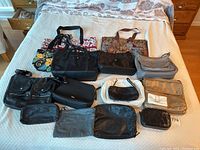 Wide view of all 14 purses and bags laid out on a bed showing various sizes, shapes, and colors mainly in black, gray, brown, and one white bag.