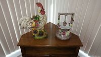 Photograph of both the pottery rooster and ceramic wishing well on a wooden surface near vertical blinds