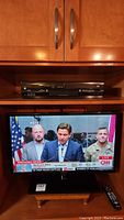 Samsung 27-inch TV powered on showing news broadcast with people in front of flags and CNN logo visible. TV is placed on a stand beneath a set of cabinets.