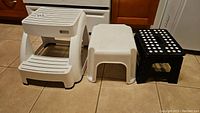 Three step stools on a tiled floor in a kitchen setting: white Mistral two-step stool, white one-step stool, and small black folding one-step stool with white polka dots.