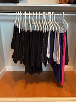 Photo of women's pants hanging on white plastic hangers on a metal bar, mostly black with some colored pairs at the end.