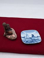 Two items on red fabric background: a blue and white ceramic dish with a mermaid and sailing boats, and a bronze mermaid figurine on a rock base, showing size and overall condition.