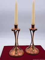Full view of the pair of Art Nouveau style copper and brass candlesticks holding taper candles on a red cloth background.