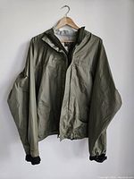 Front view of olive green Simms Gore-Tex fishing jacket hanging on wooden hanger, showing full front and left arm with elastic cuff.
