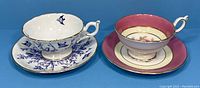 Two Coalport teacups with matching saucers against a blue background, showing blue floral and bird design set and pink floral design set.