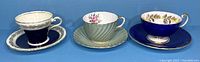 Three Aynsley teacups and matching saucers displayed side by side on a blue surface, showing each unique design and color.