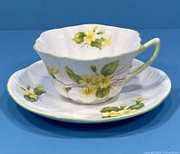 Porcelain teacup and saucer set with yellow primrose flowers and green leaves, showing front side of the teacup and saucer.