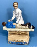 Front view of the chiropractor figurine, showing the chiropractor manipulating a patient on a wooden chiropractic table with the word 'CHIROPRACTIC' visible on the base.