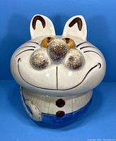 Front view of vintage cat ceramic cookie jar, showing smiling face, beige with brown accent whiskers and bright eyes, lower half painted blue resembling pants with buttons.