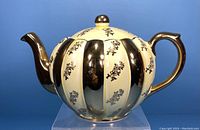 Front view of the vintage Gibson's yellow and gold ceramic teapot showing round ribbed body with alternating yellow and gold stripes and small floral pattern on yellow stripes, curved gold handle and spout, and gold knob on lid.