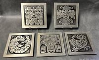 All five Native American West Coast metal plaques arranged on a black surface, showing detailed front designs of Thunderbird, Whale, Eagle, Beaver, and Bear figures.