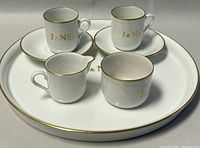 Complete 7-piece porcelain set displayed on a white surface, including serving tray, creamer, sugar bowl, two mugs and two saucers, all white with gold trim and J&NS monogram on mugs.