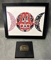 Framed Native American print with bright red, black, and purple fish design, and wooden plaque with brass emblem on dark wood base.