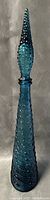 Front full view of tall blue textured glass genie bottle with pointed stopper lid.